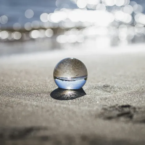 Clear glass sphere on sandy beach with blurred ocean sparkles reflecting in the background during daylight.