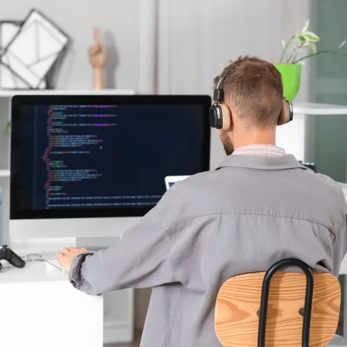 Man wearing headphones working on computer coding software at a bright, modern desk in a home office.
