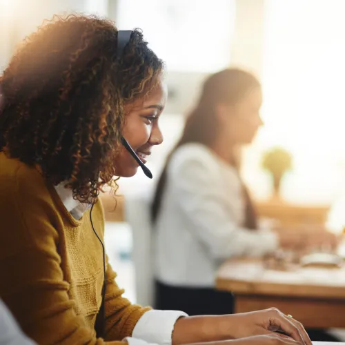 Smiling woman wearing headset working on laptop in a bright modern office with coworkers in the background