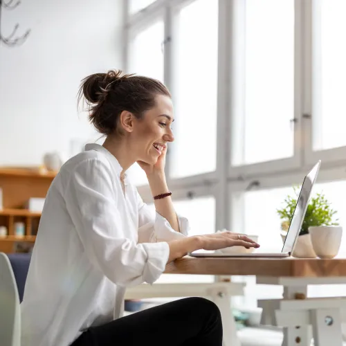 Smiling woman working on a laptop at a wooden table with a camera and potted plant near large windows.