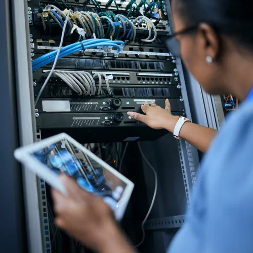 IT professional using a tablet while managing server rack connections in a data center environment