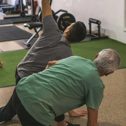 Two men stretching and exercising on mats in a gym with fitness equipment in the background.