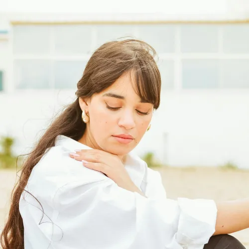 Young woman with long dark hair in white shirt and black pants sitting outdoors with eyes closed and hand on shoulder.