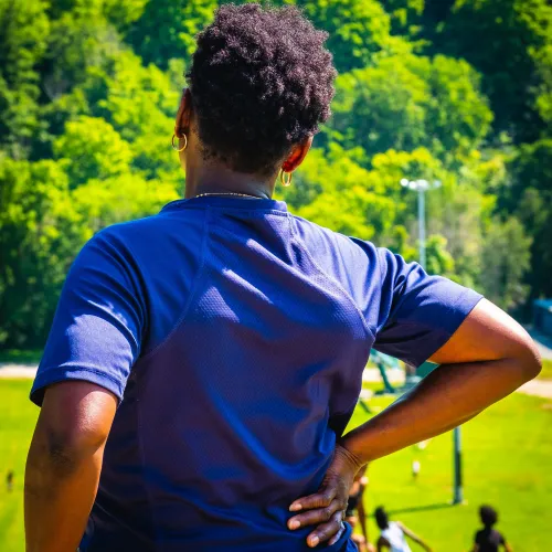 Person in blue shirt with hand on lower back overlooking a field with people playing sports surrounded by trees