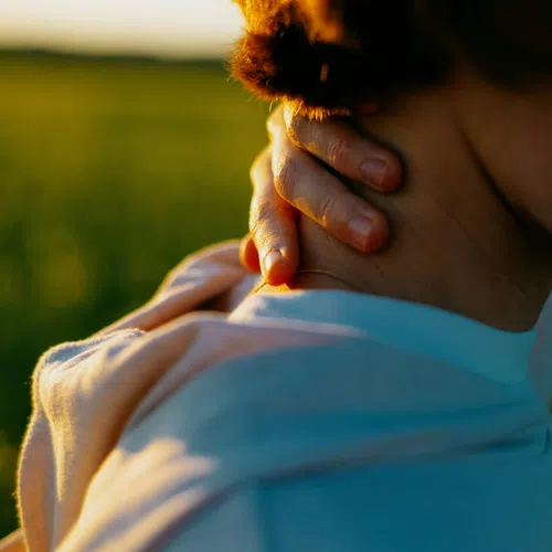 Close-up of a person in a light blue shirt holding their neck outdoors in warm sunlight.
