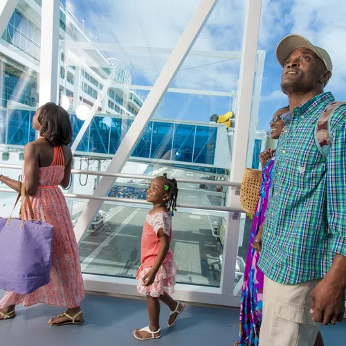 Family of four walking through a cruise terminal with a large ship visible outside the window on a sunny day