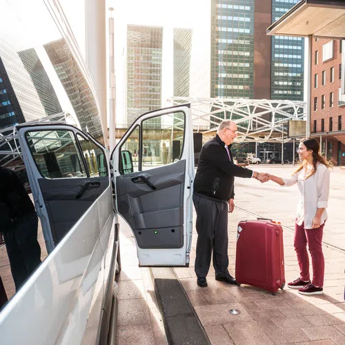 Two travelers with suitcases shake hands with a driver next to a white van in an urban setting with tall buildings.