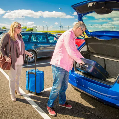 Couple loading luggage into the trunk of a blue car in a sunny parking lot with clear skies.