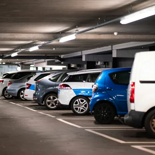 Row of various cars and vans parked in an indoor parking garage with overhead lighting and exit signs.