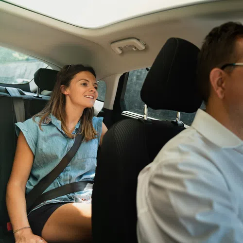 Man driving a car with a smiling woman sitting in the backseat wearing a seatbelt on a sunny day.