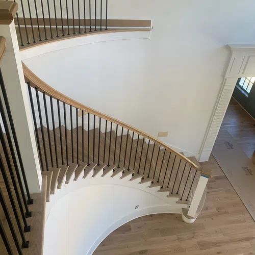 Curved wooden staircase with black iron balusters in a modern home with light hardwood floors and white walls.