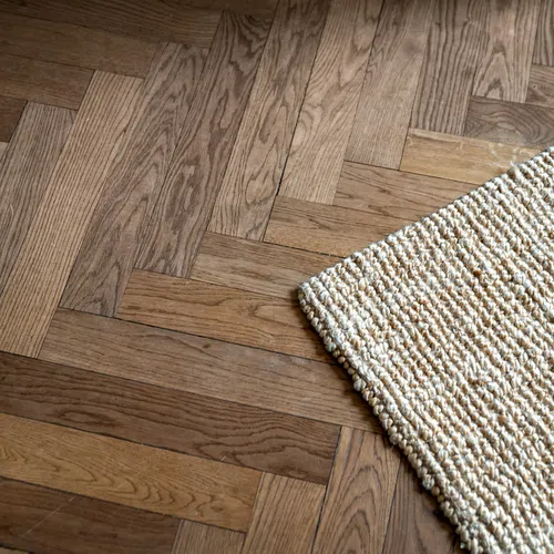 Close-up of a wooden herringbone floor with a textured beige woven rug partially visible.