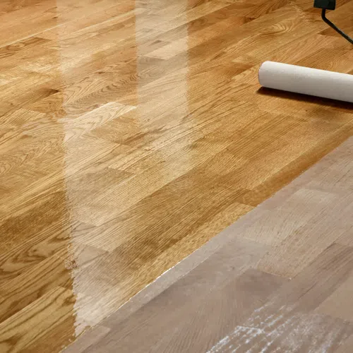 Wooden floor being polished with roller applying glossy finish showing contrast between coated and uncoated areas