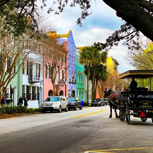 Colorful historic row houses line a street with parked cars and a horse-drawn carriage on a cloudy day.