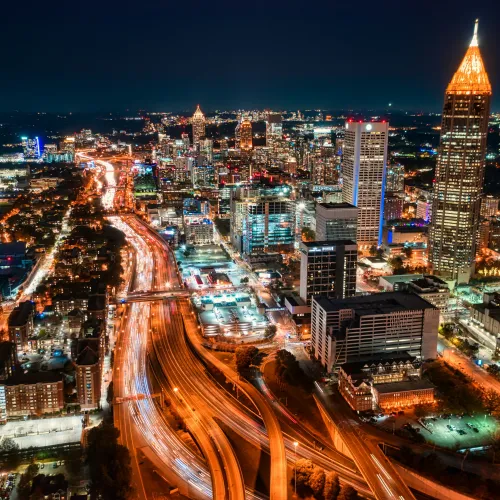 Aerial night view of a bustling city with illuminated highways and high-rise buildings including a prominent lit tower.