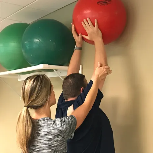 Physical therapist assisting a man reaching for a red exercise ball on a wall shelf during rehabilitation.