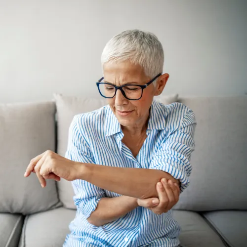 Senior woman in glasses sitting on sofa, holding and examining her painful elbow indoors with a concerned expression.