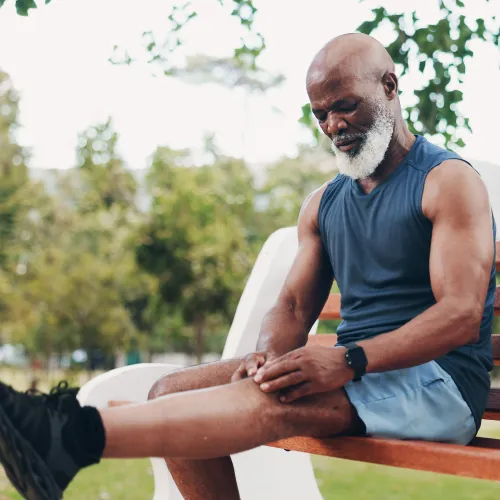 Mature man in athletic wear sitting on park bench holding his knee in pain outdoors during exercise.