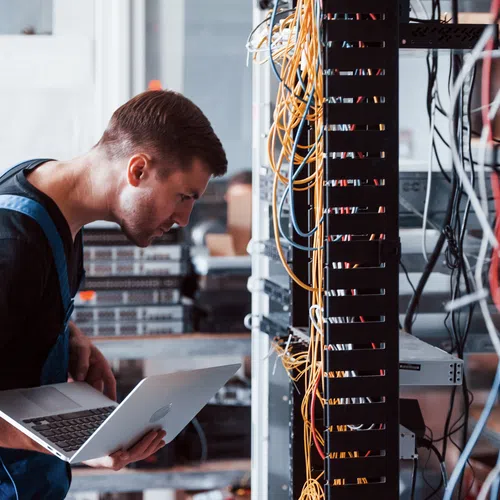 Technician in overalls inspecting server racks and cables while holding a laptop in a data center environment