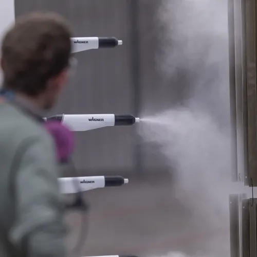 Person operating Wagner spray guns applying a mist coating to metal panels in industrial setting.