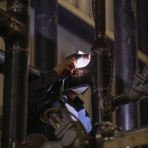 Worker welding black metal pipes in an industrial setting with welding light illuminating the area.