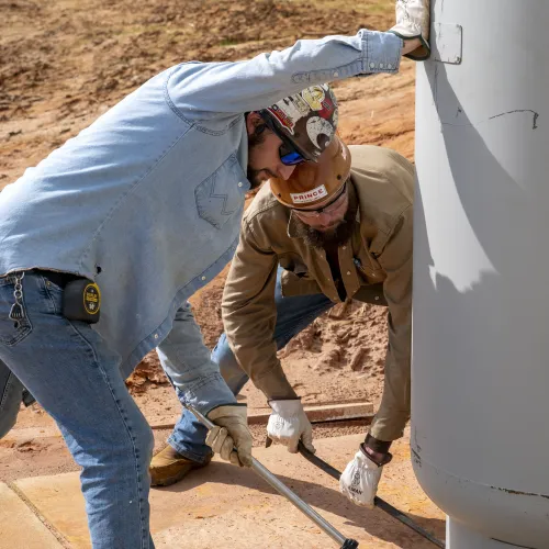 Two construction workers in safety gear working together to secure a large industrial metal tank outdoors.