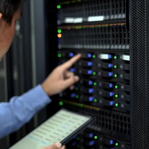 IT technician in blue shirt managing server hardware and monitoring data on a tablet in data center rack setup