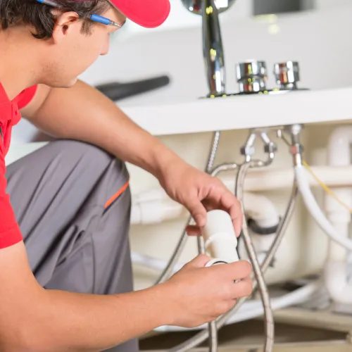 Plumber in red shirt fixing pipes under a sink, showcasing plumbing tools and water connections.