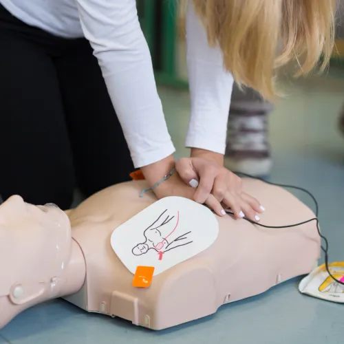 Person performing CPR training on a mannequin with an AED device nearby.