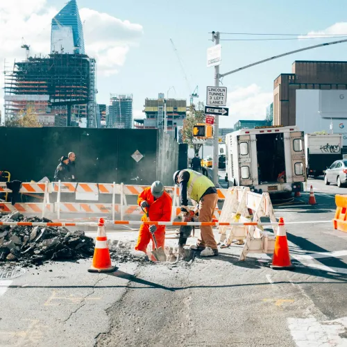 City road construction workers repairing asphalt surrounded by safety cones and barriers with urban buildings in background