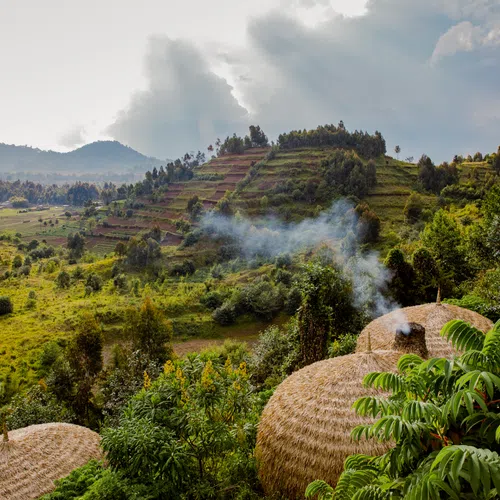 Rural landscape with thatched huts emitting smoke, green vegetation, and terraced hills under a cloudy sky.