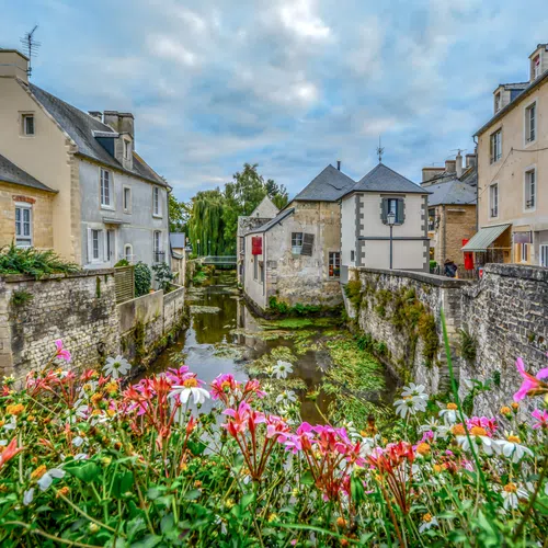 Old European village canal with stone buildings and vibrant flowers under a cloudy sky