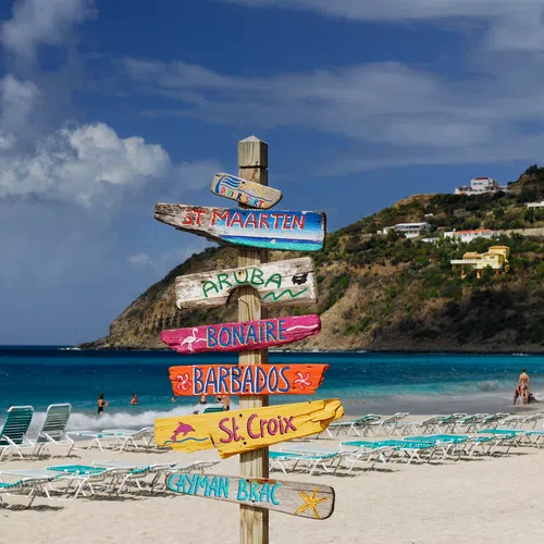 Colorful wooden signpost on a sandy beach with turquoise sea and hills in the background under a partly cloudy sky