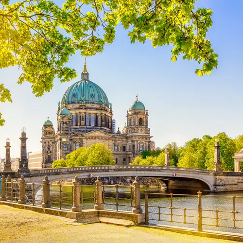 Berlin Cathedral with green dome beside a river and bridge under clear blue sky framed by spring green leaves.