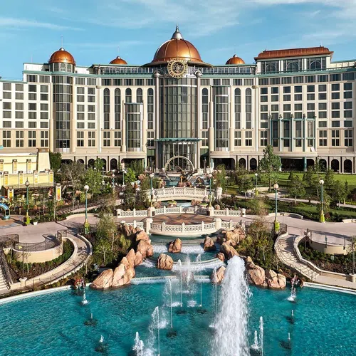 Luxurious hotel with domed rooftops overlooking a large fountain and landscaped gardens under blue sky.