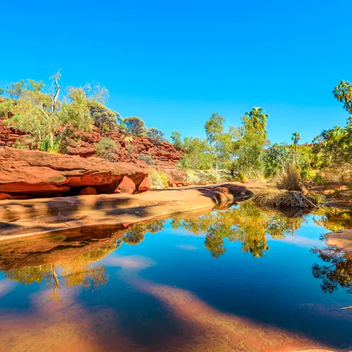 Clear blue sky over red rock formations and a calm reflective water body surrounded by green trees.