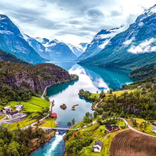 Aerial view of a fjord surrounded by snow-capped mountains, lush greenery, scattered houses, and a winding river in Norway.