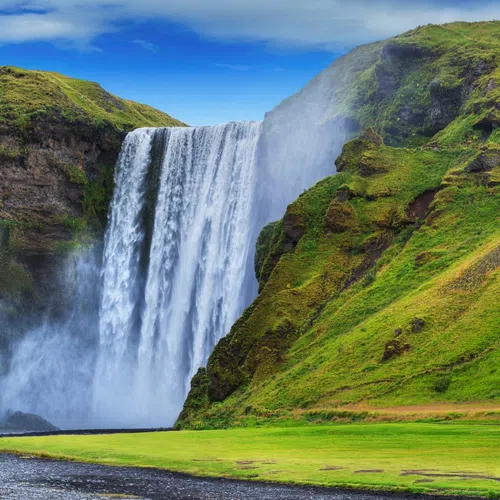 Wide view of a majestic waterfall cascading between lush green cliffs under a bright blue sky.