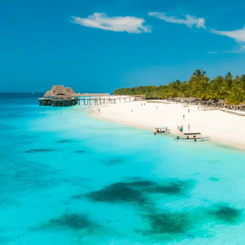 Clear turquoise ocean water meets white sandy beach with palm trees and wooden pier under a bright blue sky