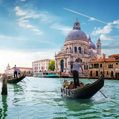 Gondoliers navigating Venice canals with Santa Maria della Salute basilica in the background under blue sky.