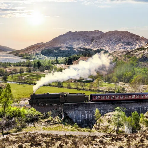 Historic steam train crossing the Glenfinnan Viaduct in scenic Scottish Highlands during sunset with hills and greenery.