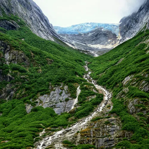 Mountain valley with lush greenery, waterfall flowing into a serene turquoise lake below cloudy sky.
