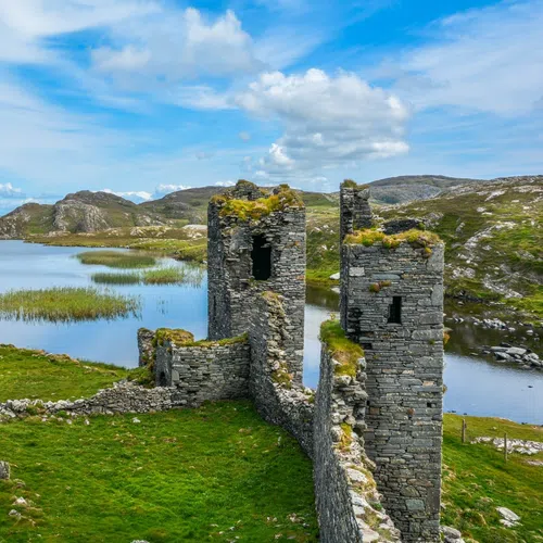 Ancient stone castle ruins beside a calm lake with grassy hills under a partly cloudy sky