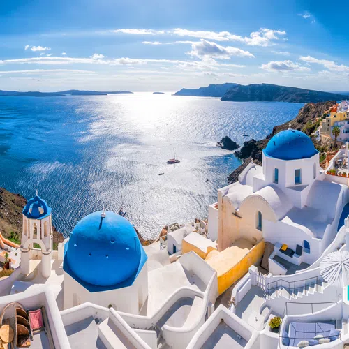 Panoramic view of Santorini's white buildings with blue domes overlooking the Aegean Sea under a bright sky