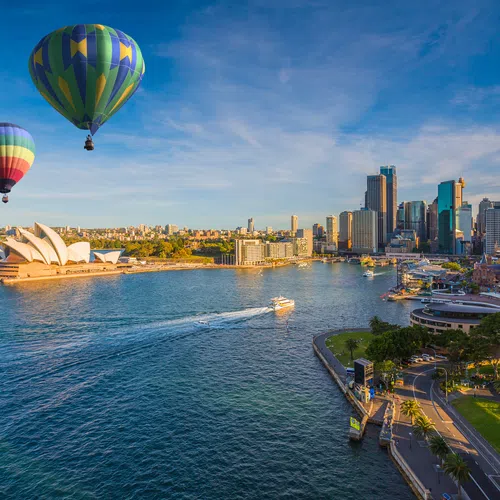 Two colorful hot air balloons floating over Sydney Harbour with the Opera House and city skyline at sunset.