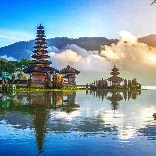 Scenic view of Balinese temple structures on a lake with mountains and sunrise in the background.