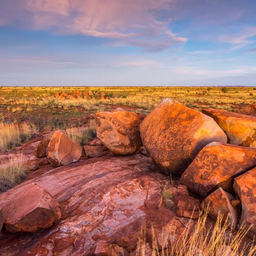 Sunlit red sandstone boulders and dry grasslands under a partly cloudy sky at sunset in the Australian outback.