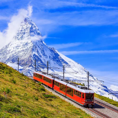 Red train climbing a steep railway with the Matterhorn mountain in background under blue sky.