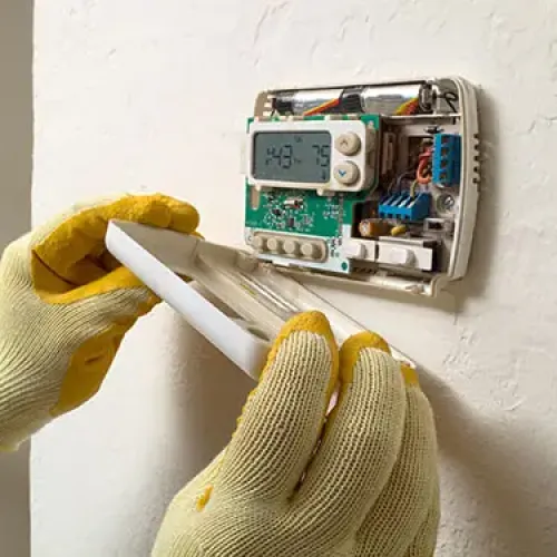 Technician wearing gloves removing cover from wall-mounted digital thermostat revealing circuit board inside.
