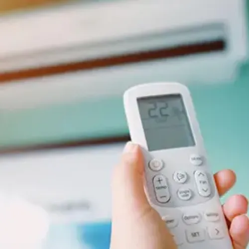 Hand holding remote controlling an air conditioner set to 22 degrees Celsius against a teal wall.